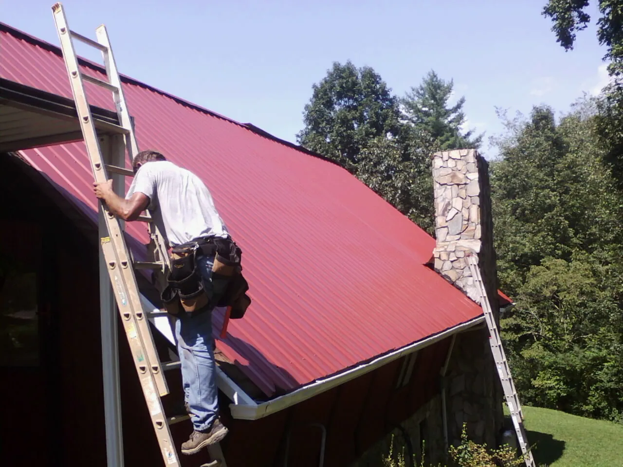 Roofer on a ladder beside a red metal roof and stone chimney — Soffit & Fascia Repair by Roofing Brothers in Pelican Rapids